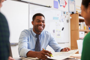 Teacher sitting at his desk smiling while talking to students.